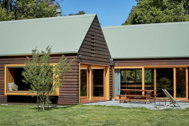 Single-storey residential home with dark horizontal timber cladding, a steep gabled central section, and broad pale green corrugated metal roofs. Warm timber-framed windows and glazed doors open onto a paved outdoor seating area with a picnic table and deck chairs. The house is set in a leafy garden with mature trees and a lawn.