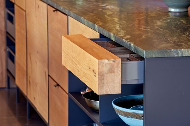 Close-up view of custom kitchen cabinetry with a large projecting timber drawer, open dark shelving, and a thick mottled stone benchtop. The warm timber fronts contrast with deep blue-grey cabinetry and ceramic bowls, highlighting detailed built-in joinery.