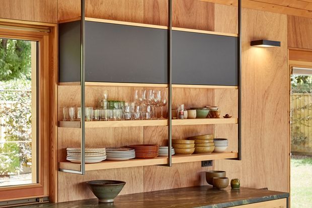 Built-in timber display and storage cabinetry with open shelving, dark inset panels, glassware, crockery and decorative bowls above a dark stone benchtop. The warm wood-lined interior includes large windows and a simple wall-mounted light, creating a refined contemporary kitchen or dining joinery feature.