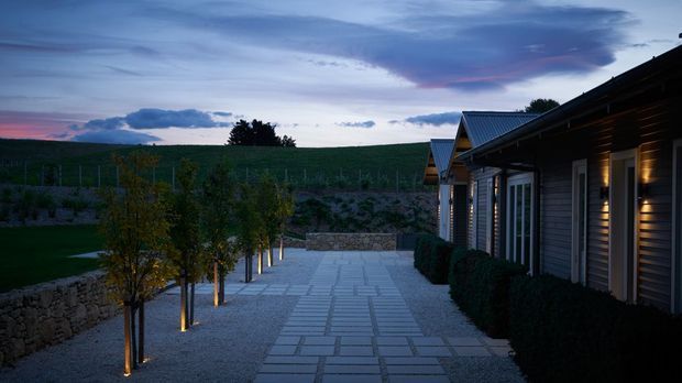 A dusk exterior view of a long, low-slung building with dark horizontal cladding, pitched metal roofs, and illuminated windows. A broad paved walkway with rectangular stone slabs runs alongside clipped hedges and regularly planted trees, with vineyard-like rows and rolling countryside visible beyond.
