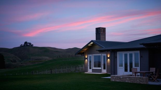 A single-storey dark-clad rural house photographed at dusk, with a pitched roof, prominent chimney, illuminated windows and entry, and a low stone-clad terrace wall. Rolling grassy hills and fencing surround the home beneath a dramatic pink and blue sunset sky.