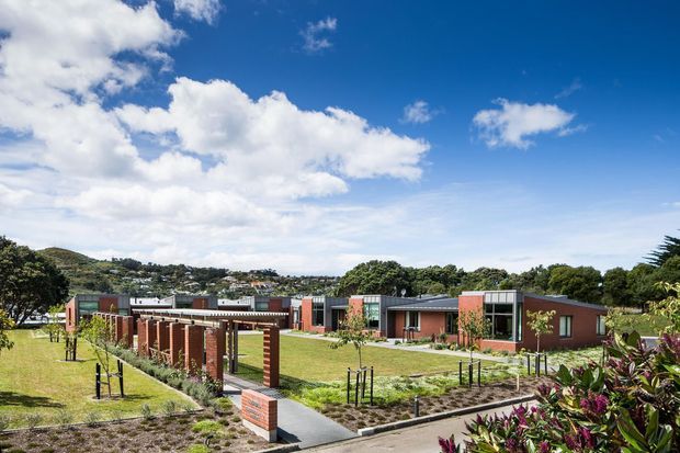 Wide exterior view of a low-rise contemporary complex set within landscaped grounds. The buildings feature flat roofs, brick-red masonry walls, extensive horizontal glazing, and dark metal framing. Paved paths, lawns, planted garden beds, and a series of brick-and-timber pergola-like screens define the site. A suburban hillside and bright blue sky form the background.