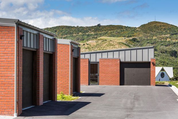 Exterior view of a contemporary low-rise brick complex with multiple dark entry doors, metal-framed clerestory-style glazing, a broad asphalt driveway, and a large dark garage door. A steep vegetated hillside forms the background, and a small white gabled structure is visible beyond the main building.