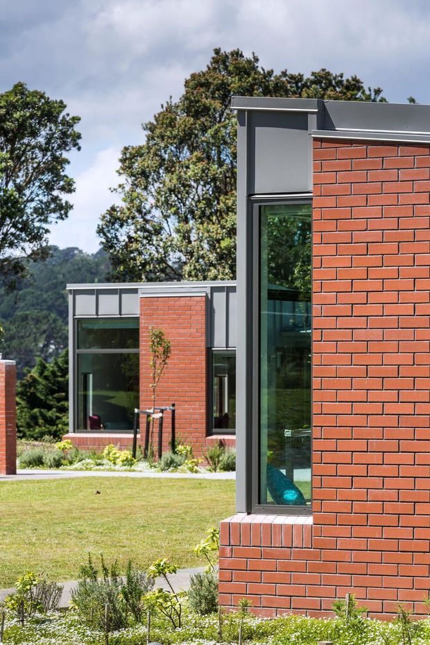 Exterior view of a contemporary low-rise building composed of red brick wall planes, dark grey metal-framed glazing, and flat rooflines. The foreground facade frames a landscaped lawn and garden, with a similar brick-and-glass wing visible behind it against a wooded hillside.