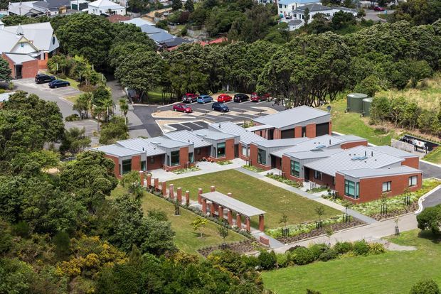 Aerial view of a low-rise contemporary brick complex arranged around landscaped courtyards and pedestrian paths. The buildings have grey metal pitched roofs, large windows, covered entry areas, and open grassy spaces, with a parking area and suburban residential neighbourhood surrounding the site.
