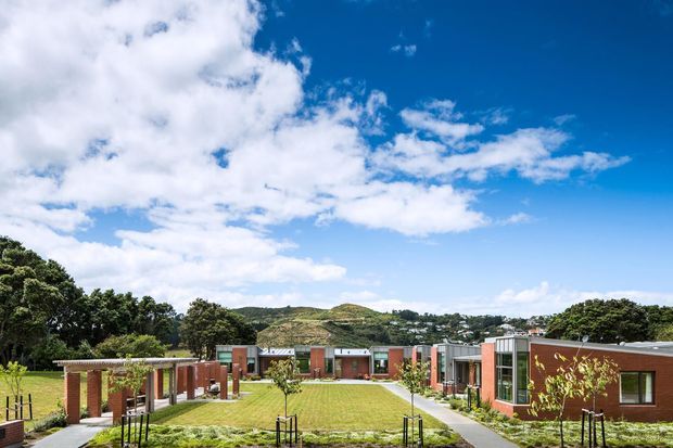 Wide exterior view of a contemporary low-rise brick complex arranged around a landscaped central lawn and pedestrian paths. The buildings feature red-brown brick walls, extensive glazing, flat or very low-pitched roofs, and modern rectilinear forms. Young planted trees, established trees, rolling hills, and a bright blue sky are visible in the surrounding setting.