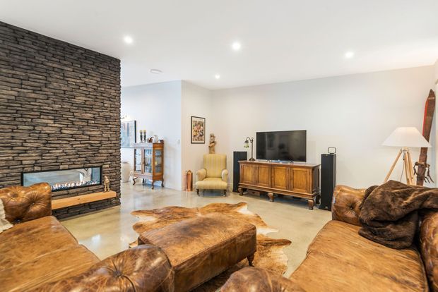 Warm contemporary living room with a prominent dark stacked-stone fireplace wall, linear gas fire, leather sofas and ottoman, cowhide rug, timber mid-century-style media cabinet, television, and a tripod floor lamp. The room opens toward a dining or kitchen area in the background.