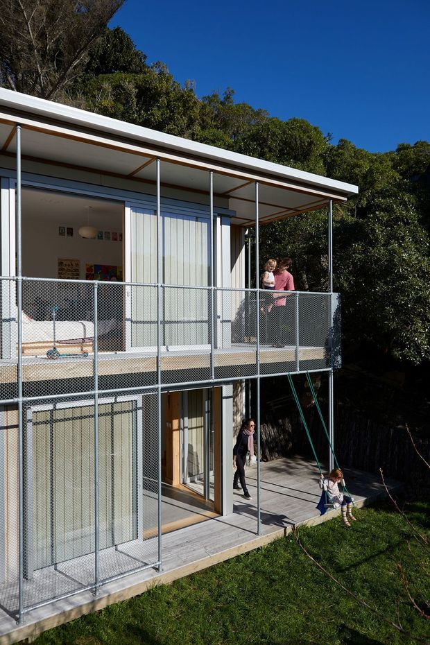 Two-storey residential home set into a leafy site, viewed from the garden. The building has a simple flat-roofed form, extensive sliding glass doors, pale vertical wall cladding, and open timber-deck balconies with galvanized chain-link safety railings. A covered upper-level terrace and lower deck are visible, with people present but not central to the architecture.