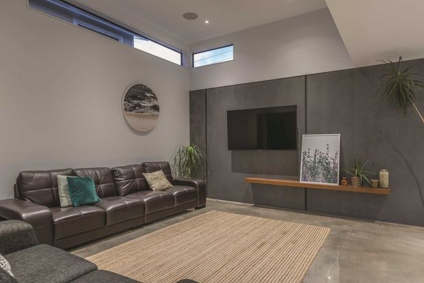 Contemporary living room with a dark leather sectional sofa, textured neutral rug, concrete-look feature wall, wall-mounted television, timber floating console, high clerestory windows, and recessed ceiling lighting. Indoor plants and restrained artwork add natural accents to the otherwise neutral, refined space.