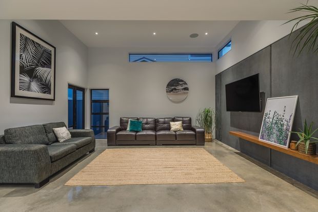 Contemporary living room with a high ceiling, clerestory windows and dark-framed glazing. The room features grey fabric and brown leather sofas, a large natural-fibre rug, polished concrete flooring, a dark concrete or stone TV feature wall with integrated timber display ledge, recessed ceiling lights, and neutral decor.