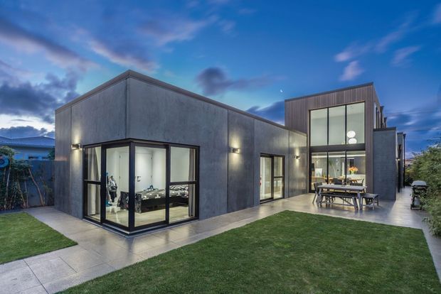 Twilight exterior view of a contemporary multi-volume residence with dark grey rendered walls, expansive black-framed glazing, and a taller double-height glazed section. The home opens onto a paved terrace with outdoor dining furniture and a neatly landscaped lawn.