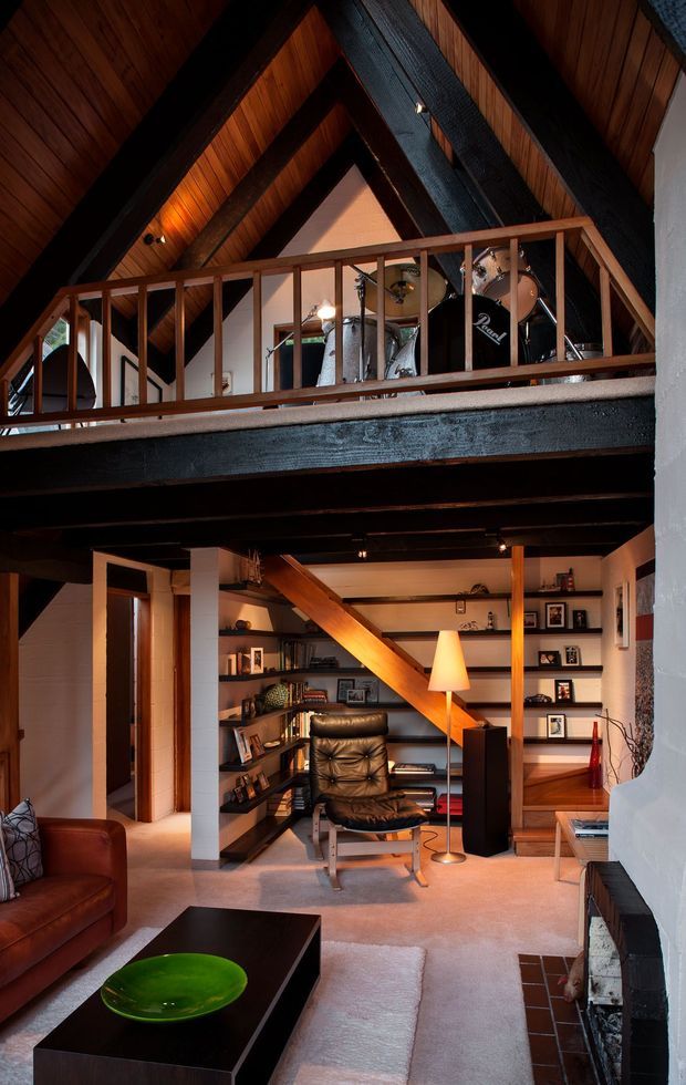 A warmly lit double-height living space with a loft mezzanine and exposed dark timber structural beams. The upper level contains a drum kit behind a timber balustrade, while the lower level features built-in open shelving, a leather lounge chair, floor lamp, sofa, coffee table and fireplace. The steeply pitched timber-lined ceiling and extensive timber joinery create a rustic, cabin-like atmosphere.