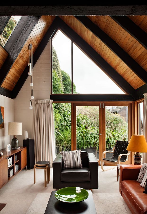 A warm, richly detailed living room with a dramatic vaulted timber ceiling and exposed dark structural beams. A large triangular window and glazed timber-framed doors bring in abundant natural light and overlook dense greenery. The room includes a black leather armchair, brown leather sofa, patterned cushions, a woven lounge chair, timber sideboard, lamps, a black coffee table and a green decorative bowl, creating a refined mid-century-inspired interior.