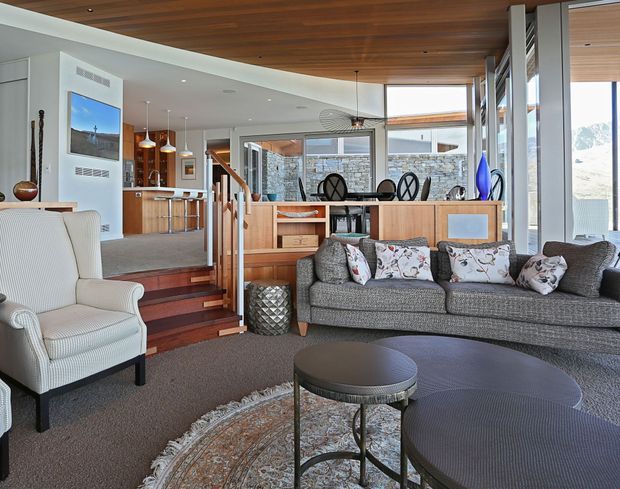Open-plan living area with a grey sectional sofa, armchair, round coffee tables and patterned rug in the foreground. A timber-lined ceiling extends through the space, with timber built-in shelving and cabinetry separating the living room from the dining area and kitchen. The kitchen includes a timber island with pendant lights, while stairs lead to a raised dining zone. Large windows and glazed doors provide abundant natural light, and a stone exterior wall is visible beyond the glazing.