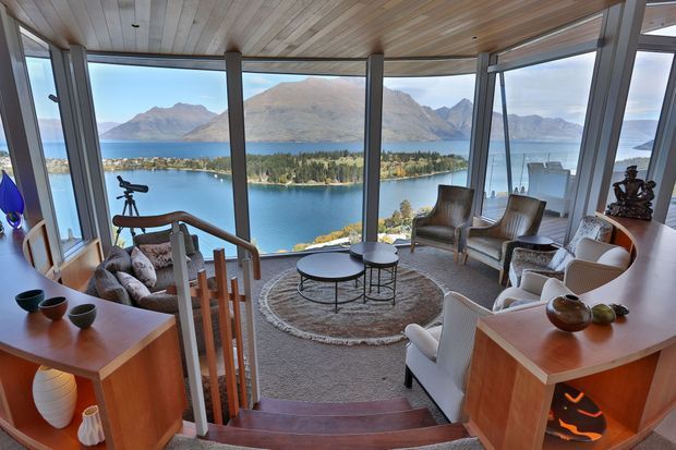 A warm, wood-lined living room or sunroom with expansive floor-to-ceiling glazing overlooking a dramatic lake and mountainous landscape. The room is arranged around a round coffee table with upholstered armchairs and sofas, with timber cabinetry and a short stairway leading down into the seating area. Natural light, panoramic views, and layered neutral furnishings create an inviting, serene atmosphere.