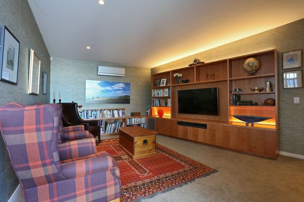 A cozy living room featuring a built-in timber media wall with television, bookshelves, display niches, cabinetry, and warm integrated lighting. Two large plaid upholstered armchairs face the entertainment unit across layered patterned rugs and a wooden storage chest. Textured grey-green walls, framed artwork, recessed ceiling lights, and a wall-mounted split-system air conditioner complete the room.