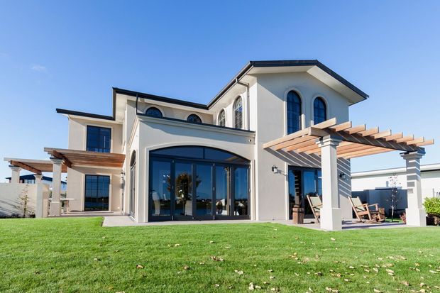 A large two-storey Mediterranean-inspired residence viewed from the landscaped lawn. The pale stucco facade features dark-framed arched windows and doors, a low-pitched multi-section roof, broad glazing, and timber pergolas over outdoor entertaining areas. The setting includes a manicured lawn, paved patio, and outdoor seating.
