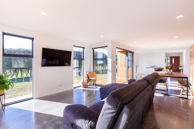 Bright contemporary open-plan living and dining area with a large dark sectional sofa, wall-mounted television, timber dining table, and extensive floor-to-ceiling glazing opening toward an outdoor area. The room has white walls, recessed ceiling lights, dark timber-look flooring, and strong natural light.