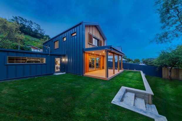 Exterior view of a contemporary multi-level home at dusk, clad in dark vertical metal panels with warm timber detailing around the upper window and covered outdoor living area. The building opens onto a manicured lawn with a paved path and built-in concrete seating or garden feature, backed by dense greenery and a sloping site.