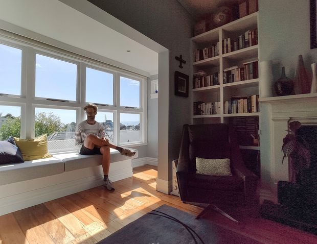 Bright, cozy living room with a built-in window seat overlooking the outdoors, a large multi-pane window, hardwood flooring, a dark upholstered armchair, full-height white bookshelves, and a traditional white fireplace with decorative shelving. Sunlight creates strong geometric shadows across the timber floor.