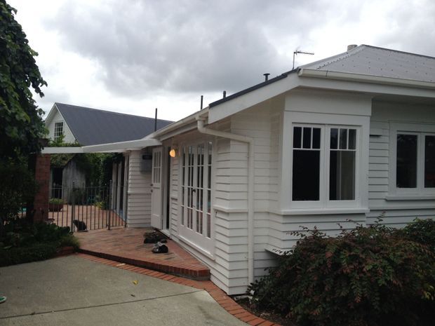 Exterior view of a single-storey residential home with white horizontal weatherboard cladding, a grey pitched roof, white-trimmed multi-pane windows and glazed French doors. A brick-paved path and small gated courtyard connect the driveway to the entry, with established garden planting around the facade.