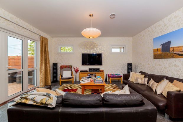 A warmly furnished living room viewed from behind a large dark leather sectional sofa. The room features patterned wallpaper, a central wooden coffee table, a colourful patterned rug, a wall-mounted television with floor-standing speakers, a sculptural pendant light, framed artwork, and full-height glazed doors opening to an outdoor deck or patio. Light enters through the glazing and small high windows, creating a cosy, eclectic atmosphere.