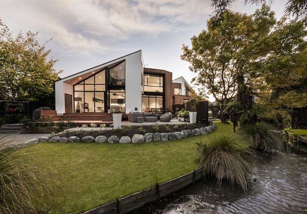 A contemporary multi-level residential house with a sharply pitched asymmetrical roof, white rendered walls, dark timber cladding, and extensive floor-to-ceiling black-framed glazing. The home opens onto a raised timber deck and landscaped lawn, with a rock-edged stream or pond in the foreground and mature planting around the site.