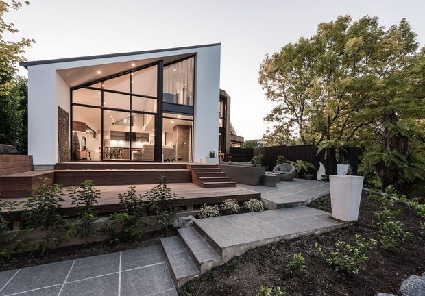 Contemporary multi-level home viewed from a landscaped outdoor area, featuring a sharply angled roofline, white rendered facade, expansive black-framed floor-to-ceiling glazing, and a warm illuminated interior. Broad timber decking and stepped concrete paving connect the garden to the living space, with raised planting beds, a dark privacy fence, outdoor seating, and mature trees surrounding the house.