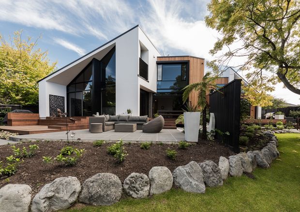 Contemporary two-storey residence viewed from a landscaped garden, featuring crisp white rendered walls, dramatic angular roof geometry, extensive dark-framed glazing, timber-clad upper-level volume, and a paved outdoor seating terrace. The front garden includes raised planting beds, large natural rocks, mature trees, and a dark vertical privacy fence.