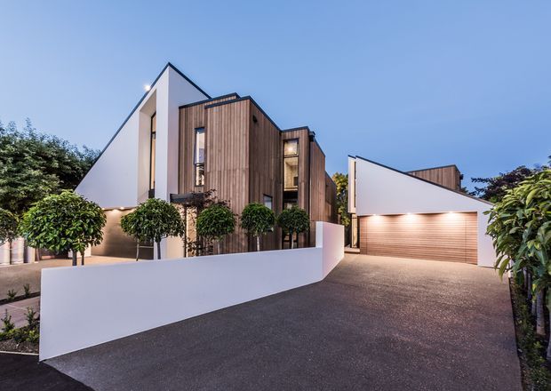 Contemporary multi-level residential exterior photographed at dusk, featuring crisp white rendered walls, vertical dark timber cladding, angular pitched roof forms, manicured topiary, a broad paved driveway, and an attached double garage with a timber-toned door.