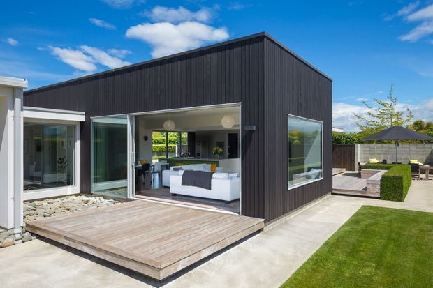 Contemporary single-storey home extension with a dark vertically clad gable-roof volume opening onto a timber deck. Large glazed sliding doors connect the interior living space to a landscaped backyard, with polished concrete paving, lawn, screening fences, and a covered outdoor dining area.