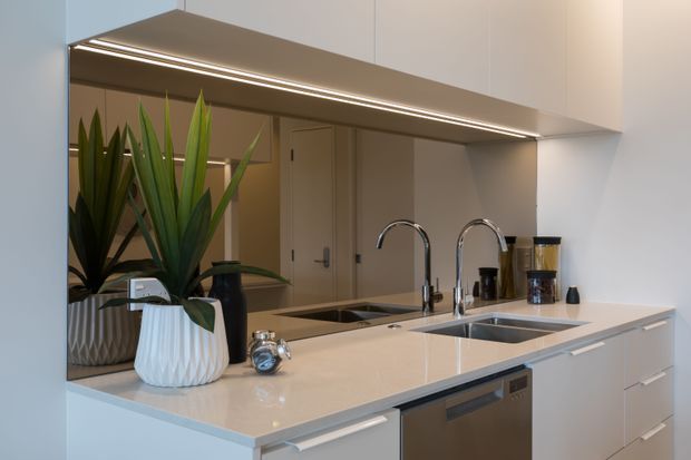 Contemporary white kitchen with a polished light stone benchtop, integrated cabinetry, double sinks, chrome gooseneck taps, and a stainless-steel dishwasher. A mirrored splashback reflects the adjacent hallway and indoor plants, while concealed linear lighting runs beneath the overhead cabinetry.