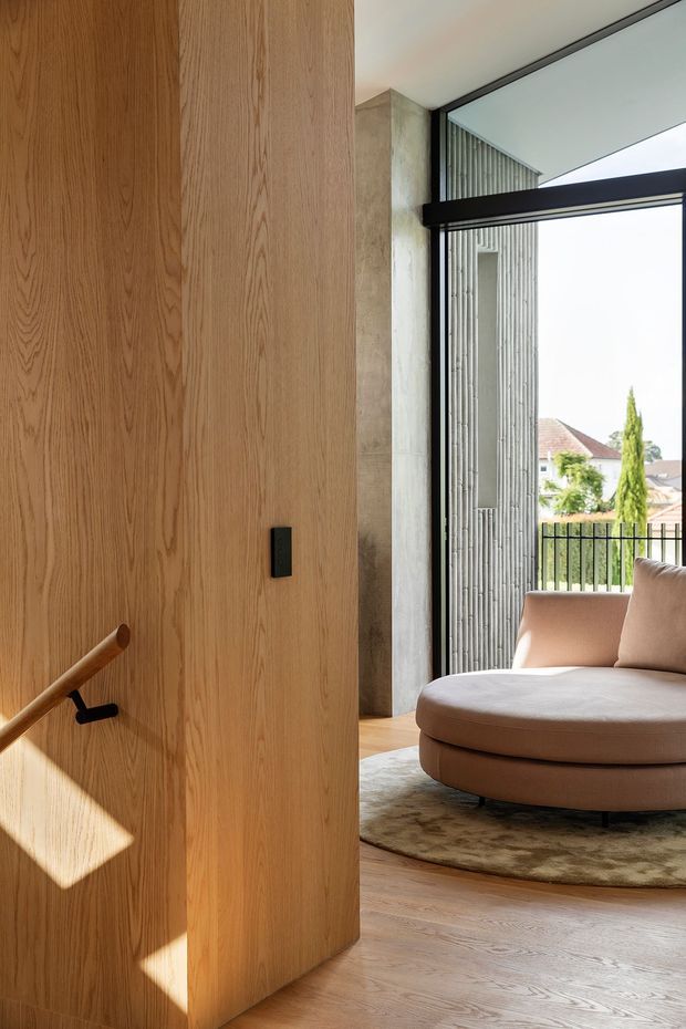A refined contemporary interior featuring a full-height light timber pivot door with a slim black handle, opening toward a sunlit sitting area. The room includes warm timber flooring, a rounded upholstered lounge chair in a muted blush-beige tone, a low-pile organic-shaped rug, dark-framed floor-to-ceiling glazing, and a ribbed concrete or masonry privacy screen outside. Strong natural light creates geometric shadows across the timber door and floor.