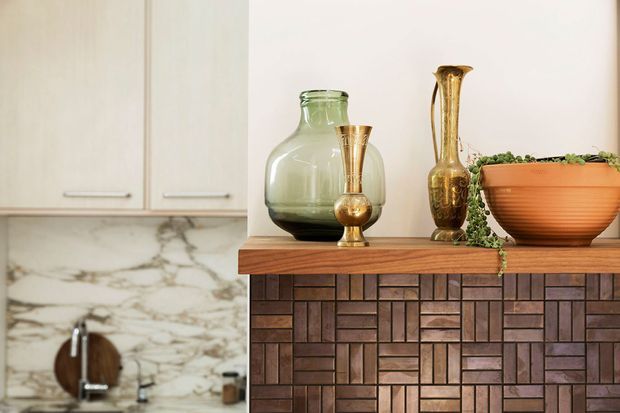 Close-up interior view of a kitchen or dining area featuring a dark timber mosaic-faced built-in cabinet or island in the foreground, topped with decorative glass and brass vessels, a terracotta bowl with trailing greenery, and a thick timber benchtop. In the background are pale flat-panel kitchen cabinets, a dramatic veined stone splashback, and a sink area.
