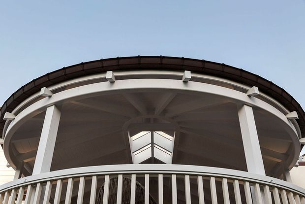 Upward-looking architectural view of a grand curved portico or balcony, featuring white classical columns, a prominent balustrade, a deep overhanging curved roof, and a central skylight or clerestory opening. The composition emphasizes the building’s ornate formal facade and strong radial ceiling geometry.
