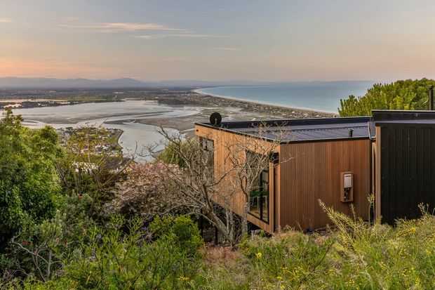 A modern dark-clad home is set into a vegetated hillside overlooking a broad coastal estuary, sandy shoreline, and open sea. The building has a low-profile flat roof, vertical warm timber cladding, dark metal cladding, and expansive glazing, with dense native-looking planting in the foreground.
