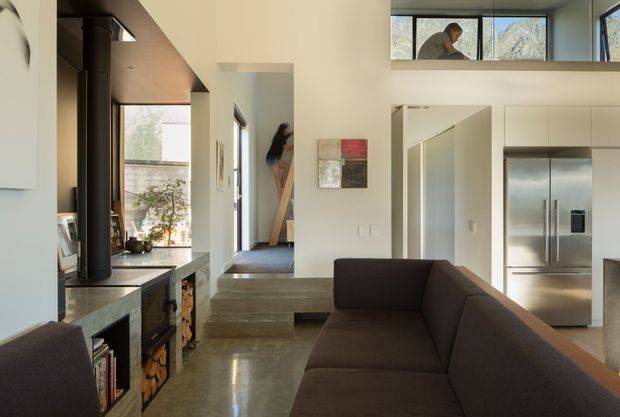 Open-plan contemporary residential interior with a living room in the foreground, a dark sectional sofa, polished concrete flooring, and a compact fireplace or wood-burning stove integrated into a dark built-in joinery wall. A short set of concrete steps leads to a raised hallway or entry area, with a stainless-steel refrigerator visible in the adjoining kitchen zone. High clerestory windows and expansive glazing bring in natural light, while a person is visible through an upper window.