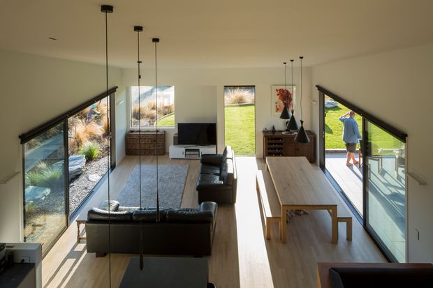 Bright contemporary open-plan living and dining room with full-height corner glazing, light timber flooring, black leather sofas, a long timber dining table, a compact media unit, and multiple slender pendant lights. The room opens directly onto a green outdoor area and is defined by strong linear glazing and a pitched or angled window-wall composition.