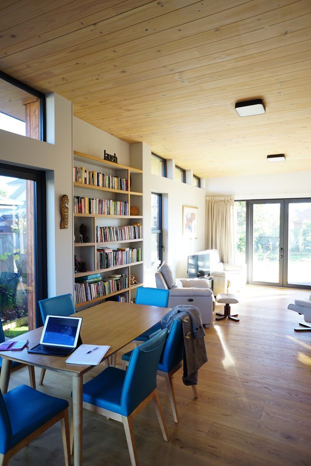 A bright open-plan living and dining space with a timber-lined ceiling, light timber flooring, built-in shelving filled with books, and large black-framed windows and glazed doors. A timber dining table with blue upholstered chairs occupies the foreground, while a relaxed seating area sits beyond. The room has abundant natural light and a warm, contemporary character.