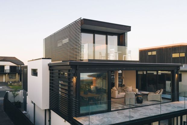 Contemporary multi-level residential building with a flat roof, dark horizontal slatted cladding, white rendered walls, extensive floor-to-ceiling glazing, and a rooftop terrace enclosed by clear glass balustrades. The image shows a substantial outdoor terrace adjoining a refined living area, with neighbouring homes visible in the background.