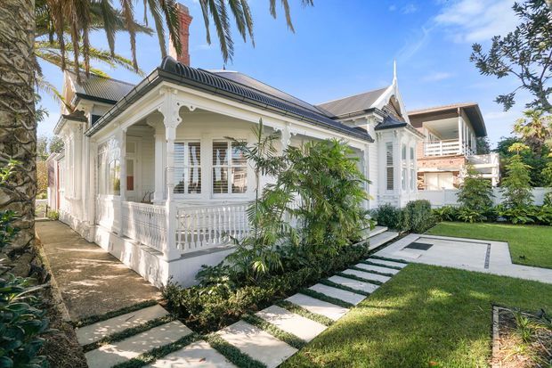 A renovated white timber villa with ornate fretwork, a wraparound front veranda, decorative columns and balustrades, and a dark pitched roof. A landscaped garden and stepping-stone pathway lead to the entrance, with a contemporary multi-level building visible behind it.