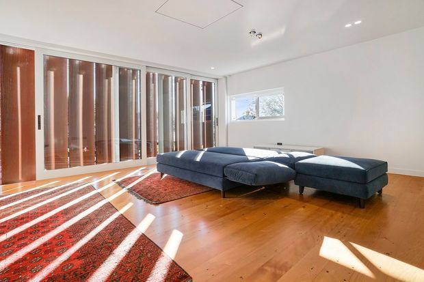 Bright, spacious living room with a large dark blue sectional sofa on a patterned red rug. The room has warm timber flooring, white walls and ceiling, extensive timber-framed glazing or sliding doors with vertical blinds, and strong sunlight creating diagonal shadows across the floor. A small high window and recessed ceiling lighting are also visible.