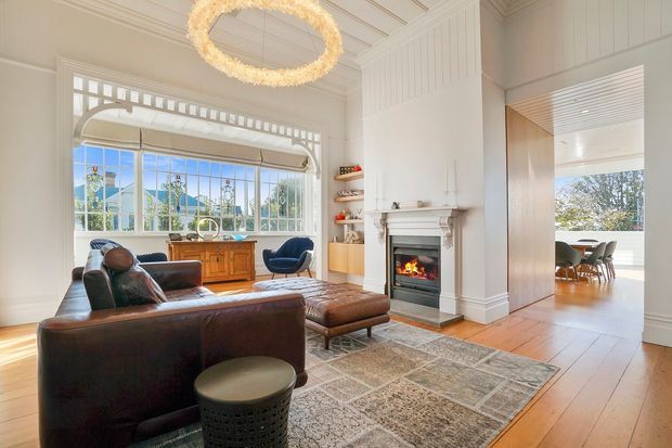 Bright, refined living room with high decorative timber-lined ceilings, white wall panelling, a traditional fireplace with a lit fire, and a large bay-style window with patterned glazing. A dark leather sofa and ottoman sit on a layered neutral rug, while an open connection leads to a dining area with a timber table and upholstered chairs. The room combines heritage architectural detailing with contemporary furnishings.