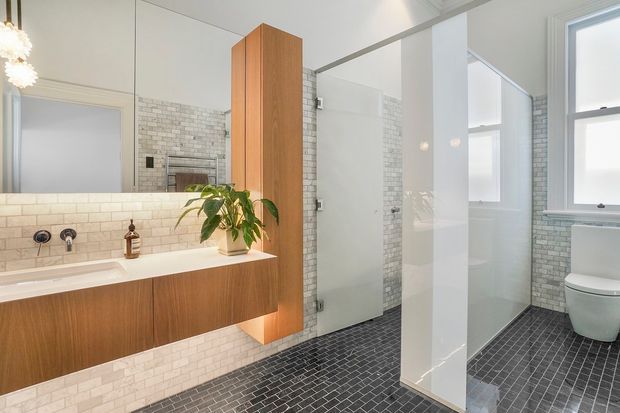 Bright contemporary bathroom with a floating timber vanity, white solid-surface benchtop, wall-mounted basin, large mirror, glass-enclosed shower, freestanding toilet, and full-height white-painted partitions. White brick-look wall tiling contrasts with dark small-format floor tiling. A warm timber storage cabinet and indoor plant add natural texture.