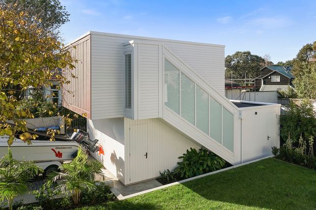 Exterior view of a compact contemporary white-clad multi-level residential building. The design features strong cubic forms, vertical and horizontal weatherboard-style cladding, a prominent glazed stair or sloping window element, and a flat roof. Landscaping and neighbouring homes surround the structure.