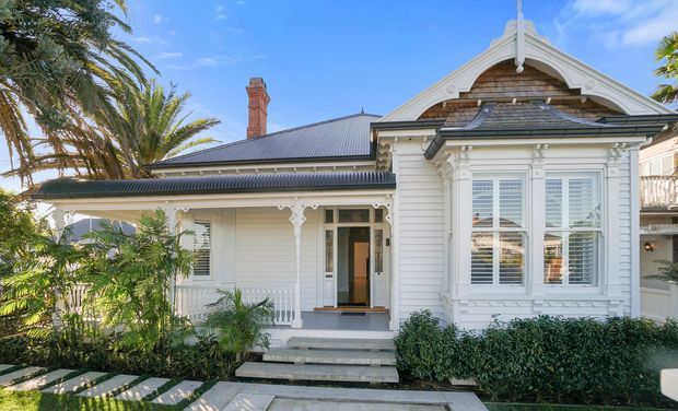 Street-facing view of a restored single-storey heritage-style cottage with white weatherboard cladding, ornate timber fretwork and gable detailing, a projecting bay window with white shutters, a corrugated metal roof, brick chimney, covered front verandah, and landscaped front garden. The image is captured from the exterior and focuses on the residential facade.