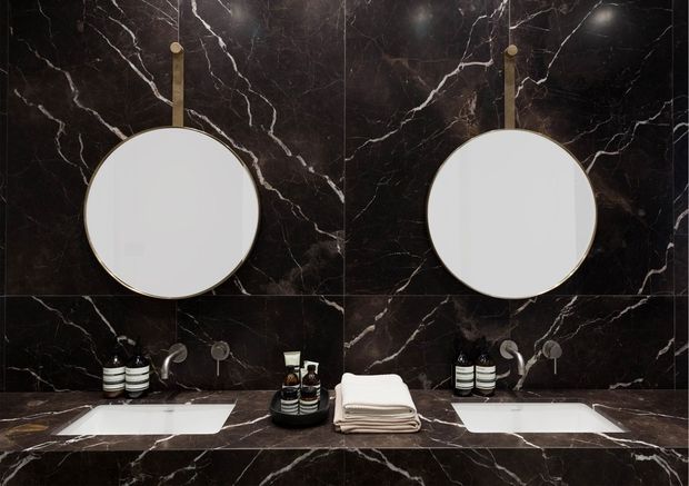 A symmetrical, luxurious bathroom vanity with two round mirrors, twin white undermount basins, and wall-to-wall dark veined marble across the walls and countertop. Brushed metal tapware, toiletries, and neatly folded towels complete the refined fit-out.