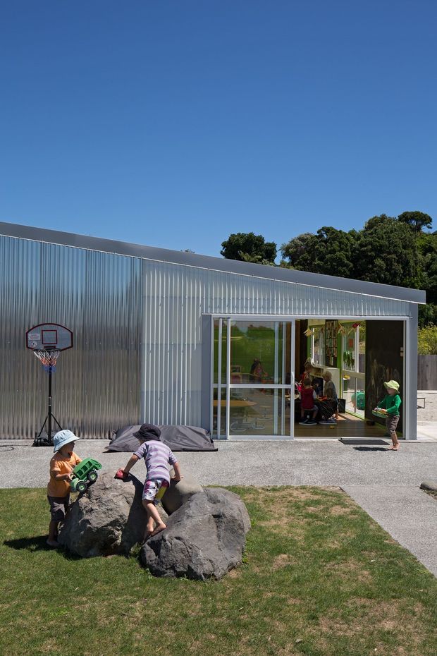 Exterior view of a contemporary single-storey recreational building with a long low roof and translucent corrugated metal wall cladding. Large glazed sliding doors open onto a paved outdoor area, with children playing in front of the building and a basketball hoop nearby. Trees and a clear blue sky are visible behind the structure.