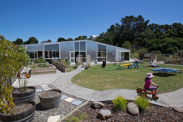 A low, single-storey contemporary building with a folded or sawtooth-like roofline, extensive floor-to-ceiling glazing, and corrugated metal cladding. It sits within a landscaped recreational grounds featuring curved gravel paths, raised planter beds, play equipment, and open lawn. The image is taken from outdoors and primarily shows the building exterior and surrounding site.