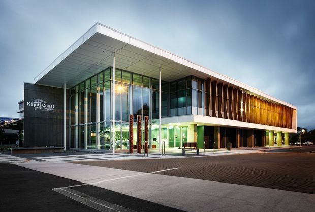 A contemporary civic or commercial building photographed at dusk. The low, elongated structure has a dramatic cantilevered flat roof, extensive floor-to-ceiling glass at the entrance, dark stone-like wall cladding, and a prominent timber-toned vertical louvre screen along the upper facade. Paved pedestrian forecourt and roadway surround the building, with illuminated interiors visible through the glazing. The sign reads “Kapiti Coast District Council.”